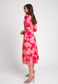 Kwiatowa sukienka w różowym materiale z czerwonymi wzorami kwiatów, z długimi bufiastymi rękawami i zwiewną spódnicą. W zestawie z beżowymi butami na obcasie.