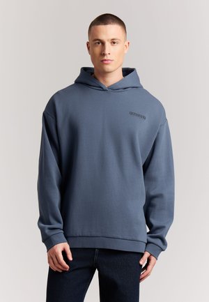 Young man with short hair wearing a blue hooded sweatshirt and dark pants standing against a plain light background.