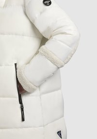 Vit pufferjacka med hög krage, stickad trim vid ärmarna och ficka med dragkedja på sidan. Mjuk, quiltad textur och logotypdetalj på ärmen.