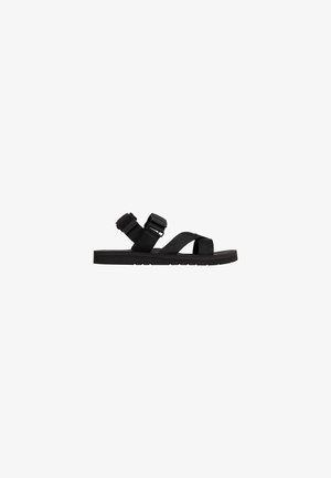 Black flat sandal with adjustable ankle and front straps, and a thick sole, isolated on white background.
