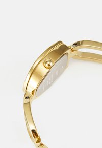 Gold-tone watch with a rounded rectangular case, textured crown, and smooth bracelet. Back features engraved details.