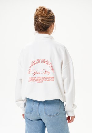 Vrouw met haar in een knot, draagt een witte trui met rode tekst "MAKE IT HAPPEN Your Way" en blauwe spijkerbroek, met de rug naar de camera op een witte achtergrond.