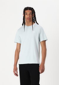 Hellblaue Baumwoll-T-Shirt mit lockerer Passform, Rundhalsausschnitt, kurzen Ärmeln und einem kleinen Logo-Detail auf der Brust. Kombiniert mit dunklen Hosen.
