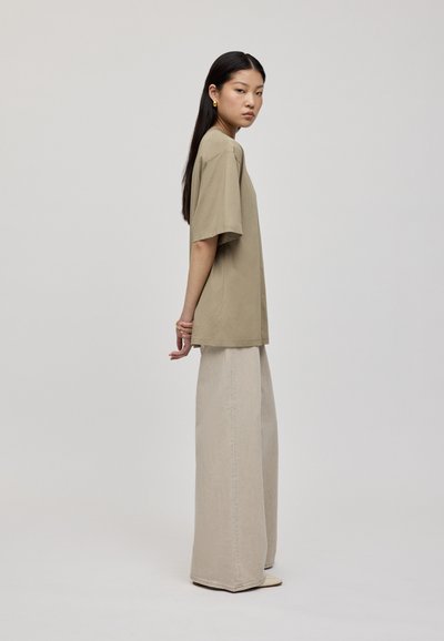 Beige oversize bavlnené tričko v kombinácii so širokými svetlo-bežovými džínsy. Model stojí bokom, predvádzajúc voľný strih a jednoduchý dizajn.