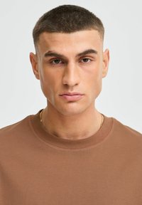 Brązowa koszulka crewneck z teksturowanej bawełny, z krótkimi rękawami i minimalistycznym designem. Model ma krótkie włosy i neutralny wyraz twarzy.