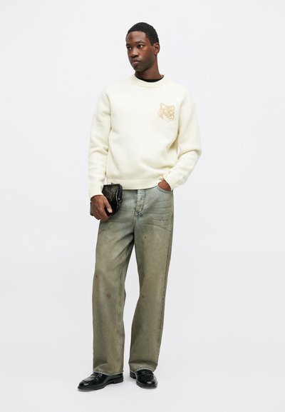 Férfi krémszínű pulóverben, állatarc mintával, laza, megfakult farmernadrágban, fekete cipőben, fekete texturált táskával a kezében, fehér háttér előtt.