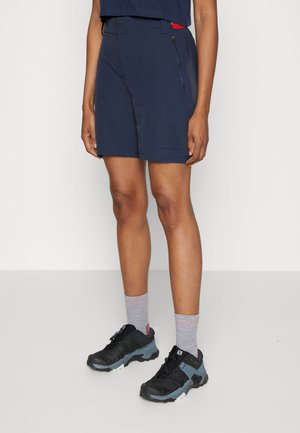 Personne portant un short bleu marine, des chaussettes grises et des chaussures de randonnée noires se tenant devant un fond blanc uni.