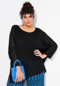 Schwarz lockeres Oberteil mit weiten Ärmeln und strukturiertem Stoff, kombiniert mit blauen, gemusterten Hosen und einer kleinen blauen Handtasche.