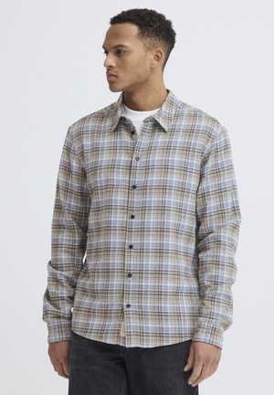 Mand iført en langærmet beige, lyseblå og grå ternet skjorte med knapper og mørke bukser, ser mod sin venstre side.