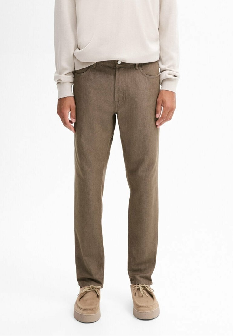 Homme portant un pantalon droit marron, un haut beige à manches longues et des chaussures en daim beiges à lacets, debout devant un fond blanc.