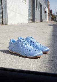 Paire de baskets en cuir bleu clair avec semelles en gomme posées sur un trottoir urbain ensoleillé près de bâtiments blancs et gris.