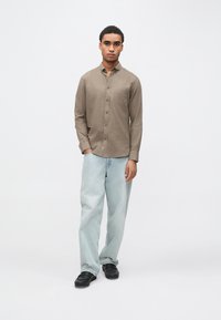 Hnedá košeľa na gombíky s dlhými rukávmi, z bavlnenej zmesi; doplnená svetlomodrými voľnými džínsami a čiernymi topánkami. Jednoduchý dizajn bez viditeľných vzorov.