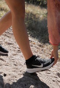 Scarpe da ginnastica nere con dettagli bianchi, tomaia in mesh con texture, suola imbottita e lacci, posizionate su terreno sabbioso.