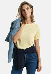 Geel katoenen t-shirt gecombineerd met een lichte spijkerjack, donkerblauwe trui rond de taille geknoopt, en blauwe spijkerbroek. Eenvoudige halslijn en pasvorm.