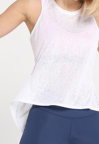 Débardeur blanc transparent avec une fente à l'ourlet et un dos légèrement allongé, présentant une texture subtile et des accents rose clair aux épaules.