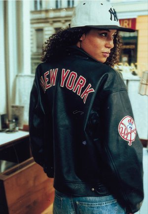 Frau mit lockigem Haar, die eine schwarze New York Yankees Lederjacke und eine weiße Yankees Cap trägt, schaut drinnen über ihre Schulter.