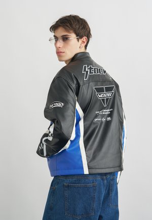 Ung mand iført en sort og blå læderjakke med hvide logoer, blå jeans og klare rektangulære briller, som kigger over sin skulder.