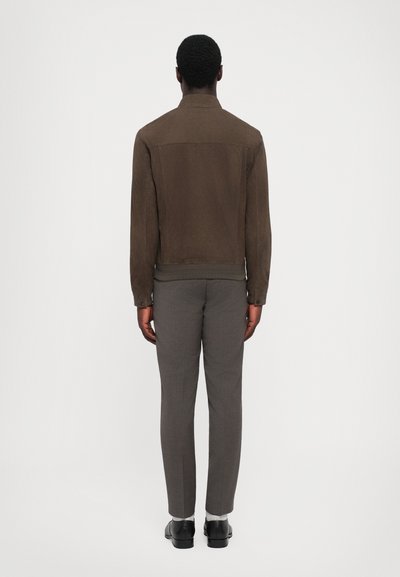 Jachetă din suede maro cu guler în picioare, design ajustat, asortată cu pantaloni din material gri slim-fit și pantofi negri. Țesătură simplă, texturată.