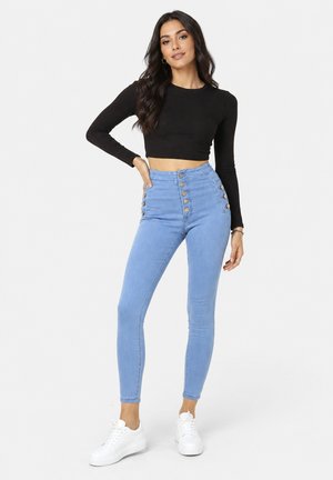 Femme portant un crop top noir à manches longues, un jean skinny bleu clair taille haute avec des boutons et des baskets blanches, debout avec la main sur la hanche.