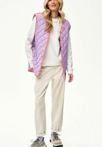 Kobieta ubrana w liliową pikowaną kamizelkę z pomarańczowymi lamówkami, białą bluzę, beżowe spodnie i szare trampki z lawendowymi skarpetkami, stojąca na białym tle.