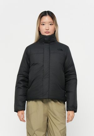 Sort dunjakke med høy krage, med horisontale sømmer og en logo på venstre bryst. Modellen har på seg lyse beige cargo-bukser.