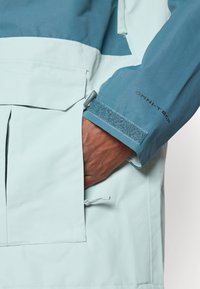 Jasnoniebieska wodoodporna kurtka z ciemniejszym niebieskim akcentem na rękawach, z rzepem przy mankietach i dużą kieszenią z przodu.