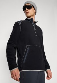 Black fleece pullover with a zippered collar, contrasting grey stitching, and side pocket; paired with a grey knit beanie and dark sunglasses.