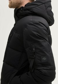 Jachetă puffer neagră cu glugă, dotată cu un buzunar pe braț cu fermoar, închideri cu buton, textură matlasată și patch cu logo pe mânecă.