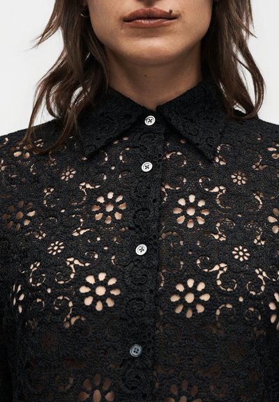 Γυναίκα που φοράει μαύρο δαντελένιο πουκάμισο με κουμπιά και ανθικό σχέδιο, με μυτερό γιακά, όπου φαίνεται το δέρμα μέσα από το δαντελωτό μοτίβο, και έχει σκουλαρίκι στο χείλος.
