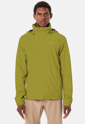 Mann trägt eine olivgrüne Regenjacke mit Kapuze und beige Hose, steht mit dem Gesicht nach vorne vor einem schlichten Hintergrund.