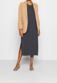 Blazer beige sopra un maxi abito blu navy con spacco laterale, abbinato a sandali con cinturini. Il blazer ha una vestibilità morbida, mentre l'abito presenta un collo rotondo.