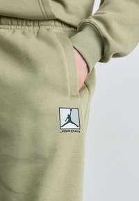 Olivengrønne joggerbukser lavet af blødt stof, med en sidelomme og et sort Jordan-logo inden for en kantet firkant på låret.