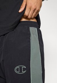 Juodi sportiniai šortai su šviesiai žalia šonine juosta, elastinga liemene ir siuvinėtu logotipu ant apatinės kojos. Audinys atrodo tekstūruotas.