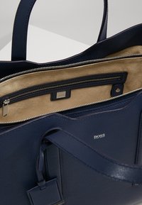 Granatowa skórzana torba BOSS z uchwytami, beżowe zamszowe wnętrze oraz wewnętrzna kieszeń na zamek błyskawiczny ze srebrnym zamkiem i plakietką z logo.