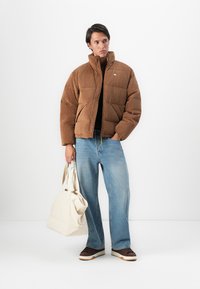 Jongeman in een bruine ribfluwelen pufferjack, lichtblauwe wijde spijkerbroek, bruine sneakers, met een grote crèmekleurige tote bag, staand tegen een witte achtergrond.