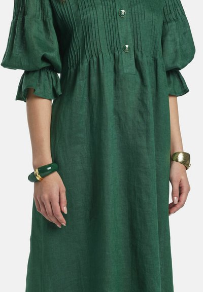 Vestido verde de lino con mangas abullonadas tres cuartos, pliegues delanteros, dos botones dorados y pulseras doradas y verdes en ambas muñecas.