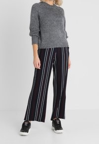 Pull gris en tricot à col rond, associé à un pantalon large noir avec des rayures verticales blanches et rouges. Des baskets noires complètent le look.
