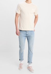 Beige bomulls t-shirt med korta ärmar och logotyp på bröstet, matchad med ljusblå jeans och rosa sneakers, stylad med vita strumpor.