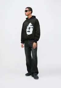 Melns kapucveida džemperis ar lielu baltu sejas grafisko attēlu, kombinēts ar tumšām džinsa biksēm un melnām slip-on kurpēm. Modelis valkā saulesbrilles.