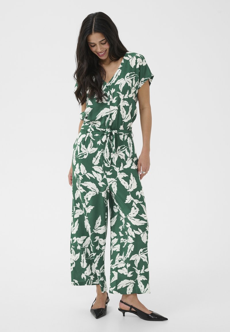 Femme souriante portant une combinaison verte avec des motifs de feuilles blanches, manches courtes, taille nouée, et des escarpins slingback noirs à bout pointu.