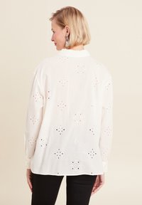 White shirt with floral embroidery and cut-out details. Features long sleeves, a collar, and a rounded hemline. Worn with black pants.