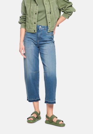 Woman wearing cropped blue jeans, olive green sandals, an olive green t-shirt, and an olive green jacket with rolled-up sleeves.