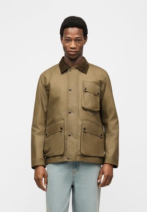 Mand stående mod en ensfarvet baggrund iført en olivengrøn jakke med sorte knapper og tre forlommer med klapper, kombineret med lyseblå jeans.