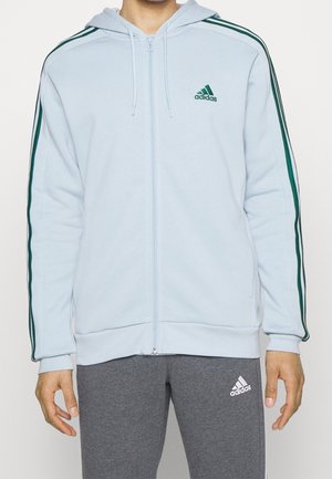 Osoba ubrana w jasnoniebieską bluzę z kapturem i zamkiem Adidas z zielonymi paskami na rękawach oraz szare spodnie dresowe Adidas, stojąca na jednolitym tle.