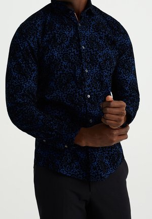 Man draagt een donkerblauw bloemenpatroon overhemd met lange mouwen, dichtgeknoopt, en stelt de manchetten bij tegen een effen lichte achtergrond.