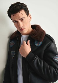 Czarna skórzana kurtka bomberka z brązowym kołnierzem z owczej wełny, z haftowanymi naszywkami w kolorze niebieskim i beżowym, z zapięciem na zamek i ściągaczami przy rękawach.