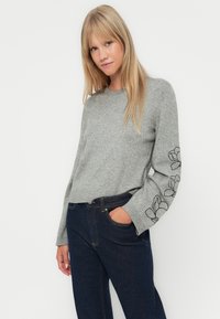 Sweater cinza de malha com decote redondo e bordados florais pretos nas mangas; combinado com jeans azul escuro. Textura suave, corte descontraído.