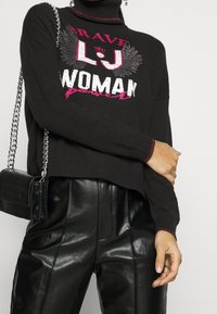 Pull noir à col montant orné de texte graphique multicolore et d'un motif d'ailes. Associé à un pantalon en cuir noir brillant et un petit sac à main.