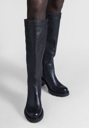 Femme portant des bottes noires en cuir texturé montant jusqu'aux genoux, avec une fermeture éclair latérale et une semelle épaisse, associées à des collants noirs transparents.