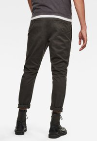 Spodnie w kolorze oliwkowej zieleni o zwężanym kroju, z mankietami na nogawkach, tylnymi kieszeniami i drobnymi detalami szwów. Noszone z ciemnymi, sznurowanymi butami.